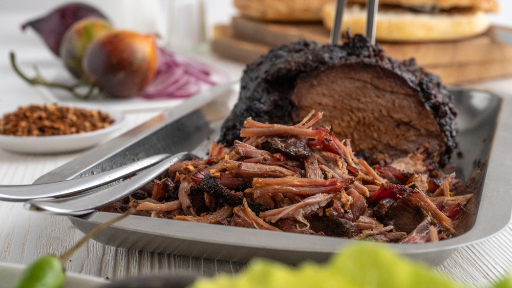 Ein Schweinebraten in einer Edelstahlschale zum Teil als Pulled Pork verarbeitet. Mit Salat im Vordergrund und Tomaten, Pul Biber und Zwiebeln im Hintergrund.