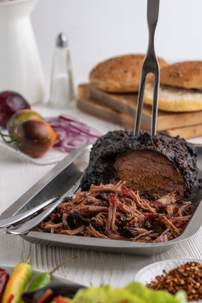 Ein Schweinebraten in einer Edelstahlschale zum Teil als Pulled Pork verarbeitet. Von oben wurde eine Fleischgabel in den Braten gesteckt. Mit Salat und Gewürzen im Vordergrund und Tomaten Zwiebeln und Dönerbrötchen im Hintergrund.