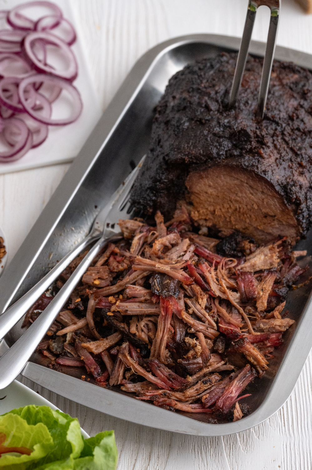 Ein Schweinbraten gegart in einer Edelstahlschale zum Teil zum Pulled-Pork verarbeitet. Von oben wurde eine Fleischgabel in den braten gesteckt. Links liegen zwei Gabeln in der Schale. Salatblätter im Vordergrund und Zwiebelringe im Hintergrund.