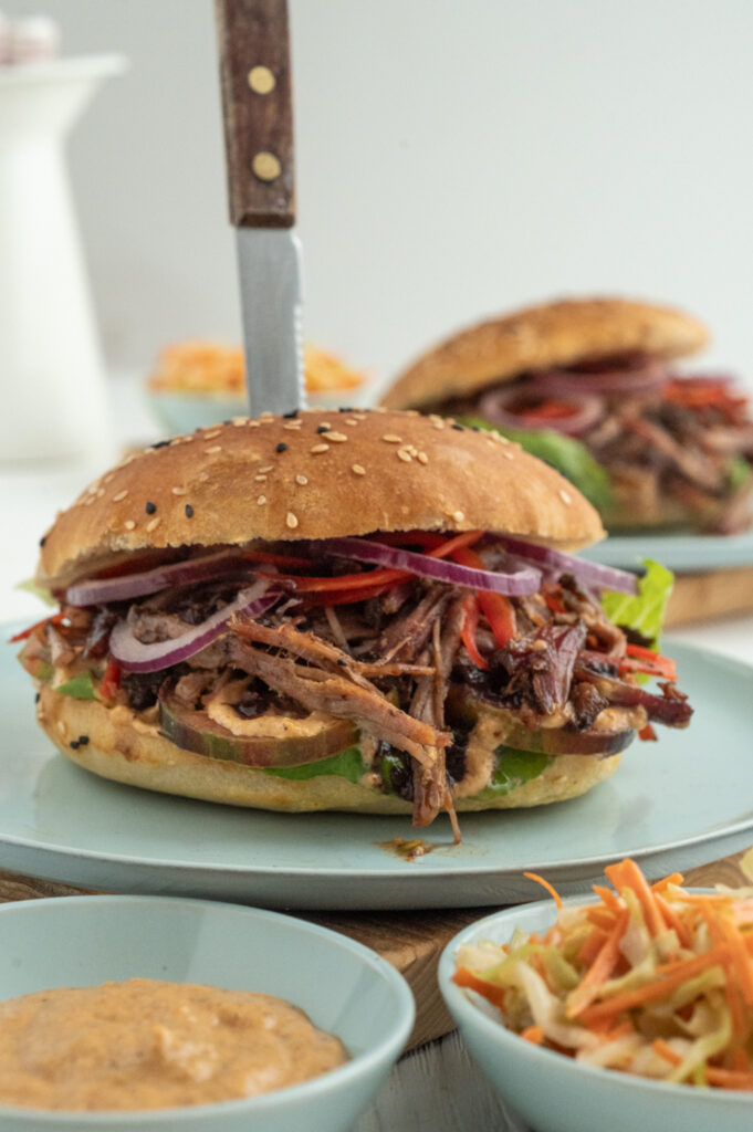 Ein Pulled Pork Burger auf einem Teller serviert. Von oben wurde ein gezacktes Messer durch den Burger gesteckt. Im Vordergrund ist jeweils in einer hellblauen Schale die Weltbeste Grill- und Burgersauce und ein Coleslaw-Amerikanischer Krautsalat mit weniger Fett und Zucker.